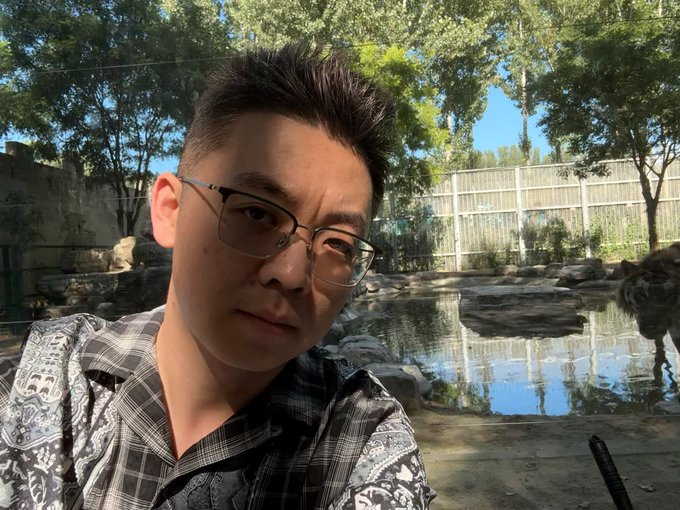 The image features a man wearing glasses, sitting near water with trees behind him. He appears to be looking at something or someone off-camera, possibly posing for a photo. The caption "卧槽有凶气" suggests that there might be some connection between the man and the location depicted in the image.
