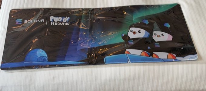 A still-sealed Pudgy Penguins branded mouse pad featuring the Solana and Pudgy Penguins logos with cartoon penguin characters against an aurora background.