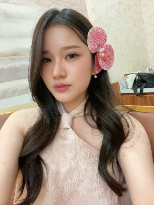 A young woman with long dark hair takes a selfie, wearing a light pink halter top and a large pink orchid flower in her hair.