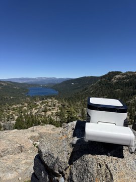 A white @maticrobots cleaning robot sits on a rocky mountain overlook, with a scenic view of a forested valley and lake stretching into the distance under a clear blue sky—capturing the novelty of taking a modern home device on an outdoor adventure.