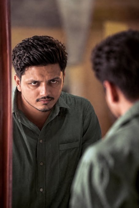 A young man with a mustache stares intensely at his reflection in a mirror, wearing an olive green button-up shirt, his expression conveying determination. The caption "GM Everyone - Another day, another trying something bogg" pairs well with the image, suggesting a mood of self-reflection and renewed motivation to tackle challenges head-on.