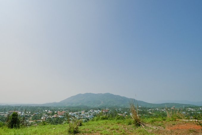 A scenic overlook of a small town nestled below a forested mountain under a clear blue sky, with tall grasses in the foreground. The image's calm, pastoral view offers a peaceful contrast to the busy crypto airdrop opportunities described in the accompanying post about Sleepagotchi, Beldex, and ETHRA SHIP campaigns.