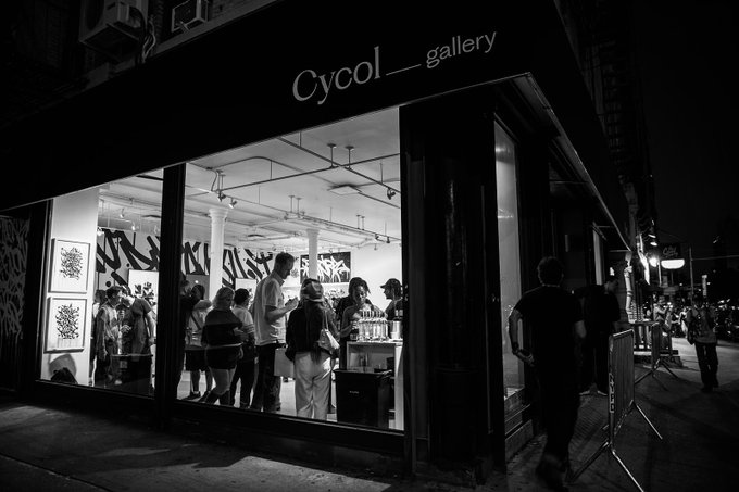 A black and white photograph captures the exterior of the 'Cycol_gallery' at night, with bright interior lights illuminating artwork on the walls and a crowd of people gathered inside, while a person walks past on the dark street outside.
