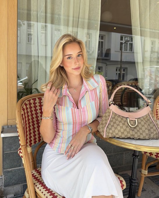 A blonde woman in a pastel striped shirt and white skirt sits at an outdoor café table with a Gucci monogram handbag featuring pink leather trim.