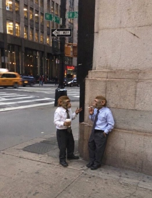 The image features two men dressed as businessmen, each wearing a tie and holding a cup of coffee while standing next to a street pole with a traffic light above them. The background includes buildings and cars, suggesting an urban setting. The caption "still the chosen one BAYC" implies that these two men are part of a special group or event, possibly related to their unique costumes.
