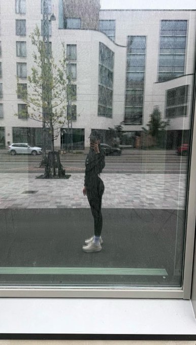 A reflection of a person in a black hoodie taking a mirror selfie with a phone, standing outside near a street with parked cars and a modern building in the background.