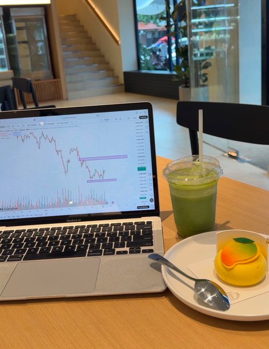 A casual workspace setup on a wooden table, featuring a MacBook Air displaying a cryptocurrency or stock trading chart, alongside a green iced matcha drink and a yellow mango mousse dessert.
