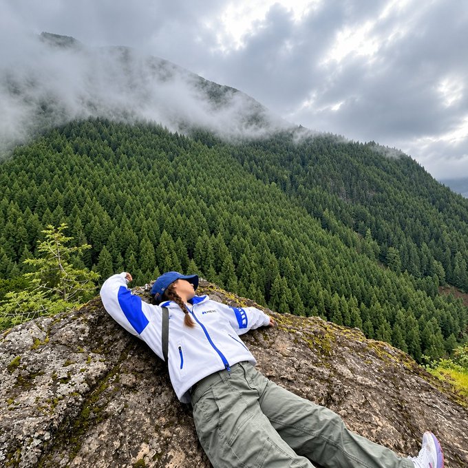 A person wearing a blue cap and white and blue jacket lying on a mossy rock, with a vast green forest and misty mountains in the background.