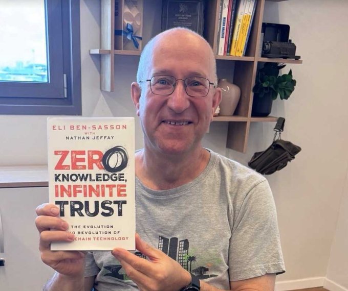 The image features a bald man holding up a book titled "Zero Knowledge, Infinite Trust" by Eli Ben-Sasson. The man appears to be smiling as he poses for the photo with the book in his hands. The background of the image shows a bookshelf filled with various books and knick-knacks, suggesting that this might be a personal library or a space dedicated to reading and learning.

The post about the book seems to be genuine and positive, as it reads "This has finally arrived! I was going to write something sarcastic about it but I can'T." The man's enthusiasm for the book is evident in his smile and the way he holds up the book.