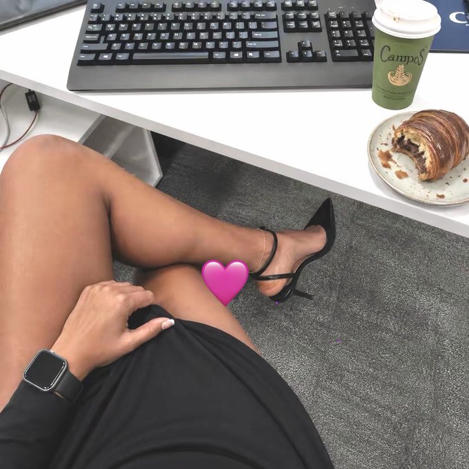 A woman in a black dress and heels relaxes under a desk while typing at a keyboard, with a "Campos" coffee cup and chocolate croissant nearby, captioned to invite others to share their favorite cryptocurrency tickers for upcoming gains.