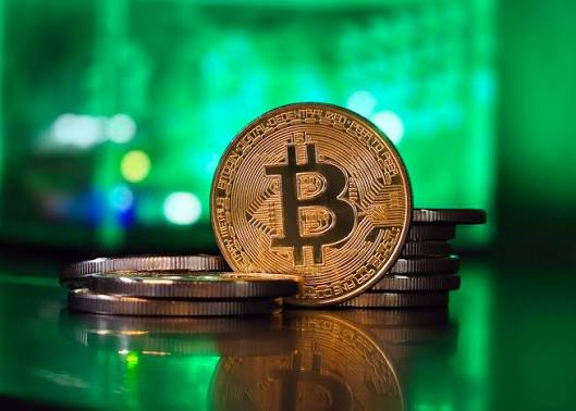 A close-up photograph of a gold Bitcoin standing upright on a stack of silver coins against a green illuminated background, with the inscription "BITCOIN DIGITAL DECENTRALIZED PEER TO PEER" visible on the coin's edge.