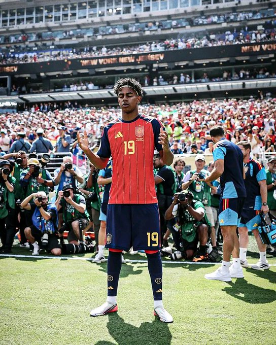Soccer player Lamine Yamal, wearing a red Spain national team jersey with number 19, stands on a sunny pitch during a 'NEW YORK NEW JERSEY' event with photographers crowded behind him.