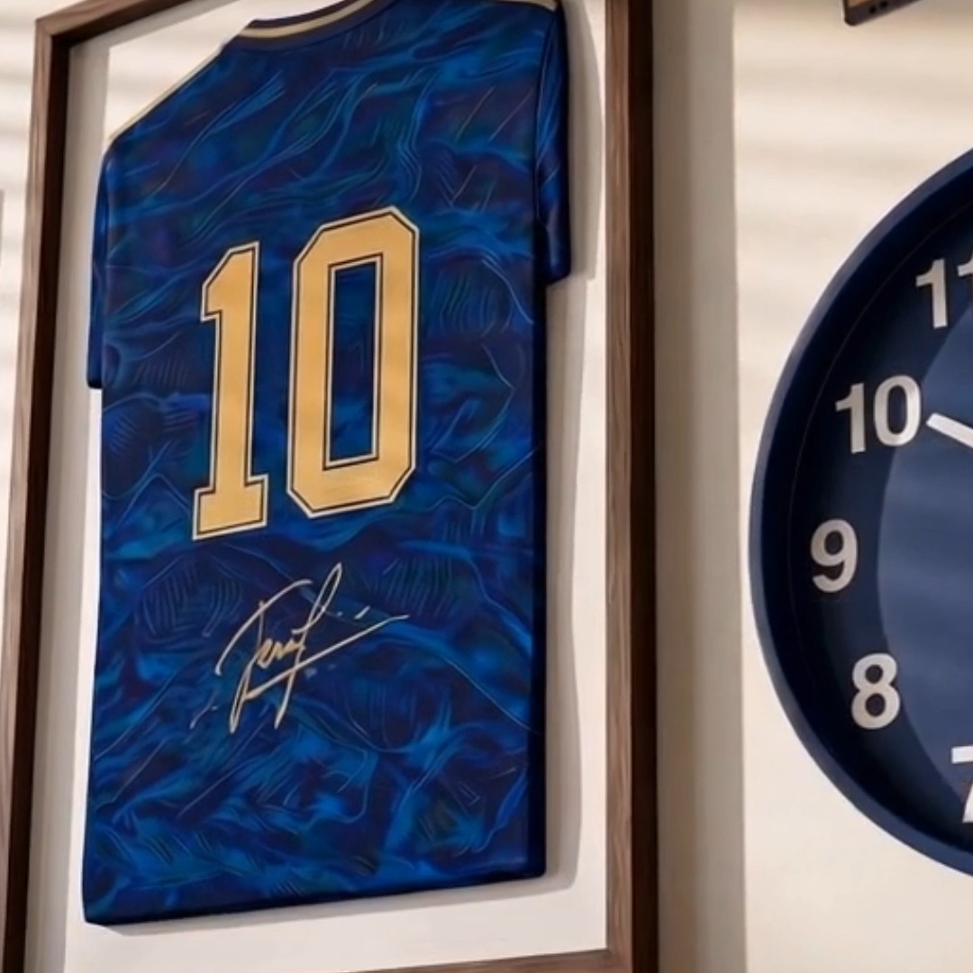 A framed blue soccer jersey with the number '10' and a signature is displayed on a wall next to a round clock.
