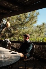 A young man with a beard wearing a dark vest sits at a glass table on a patio surrounded by pine trees, with string lights hanging above.