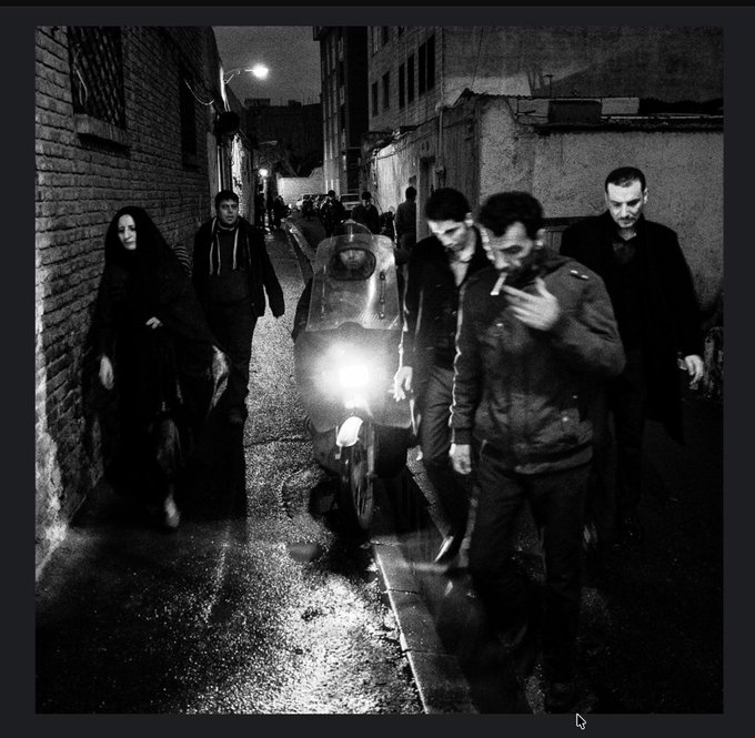 A black and white street photograph at night showing several people and a person on a scooter with a bright headlight in a narrow alleyway.