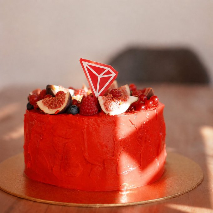 A red glazed cake topped with fresh figs, raspberries, and blueberries, featuring a red and white geometric topper resembling the TRON logo.