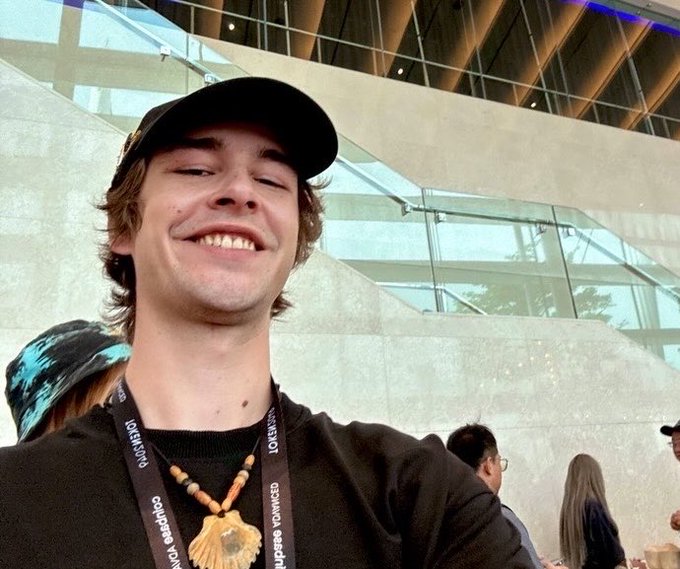 A young man wearing a black baseball cap and a shell necklace smiles at the camera in a modern building with glass and concrete architecture. The caption "Gm" likely stands for "Good morning" and relates to the image by showing someone starting their day with a positive selfie.