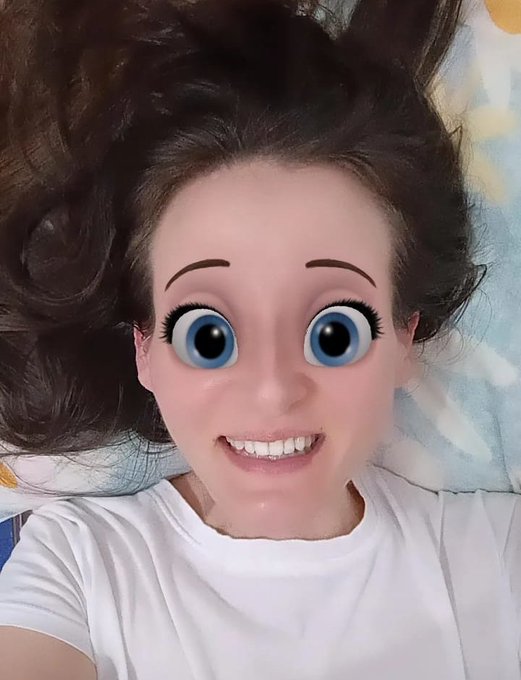 A selfie of a young woman with dark hair lying down on a floral pillow, featuring a comical cartoon face filter with oversized blue eyes and raised eyebrows.