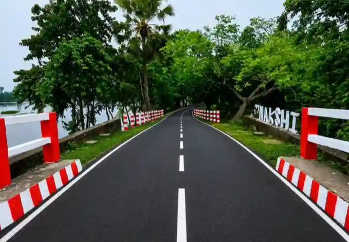 An empty asphalt road curves through lush green trees, bordered by red and white striped concrete barriers.
