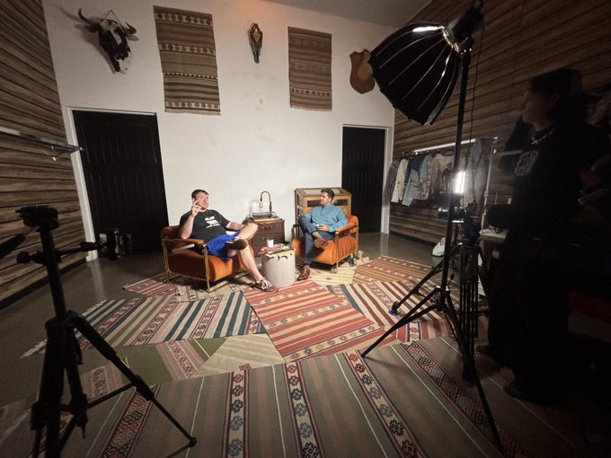 Two men sit in chairs on patterned rugs in a studio space decorated with textiles and skulls, while a crew member stands near lighting equipment filming them.