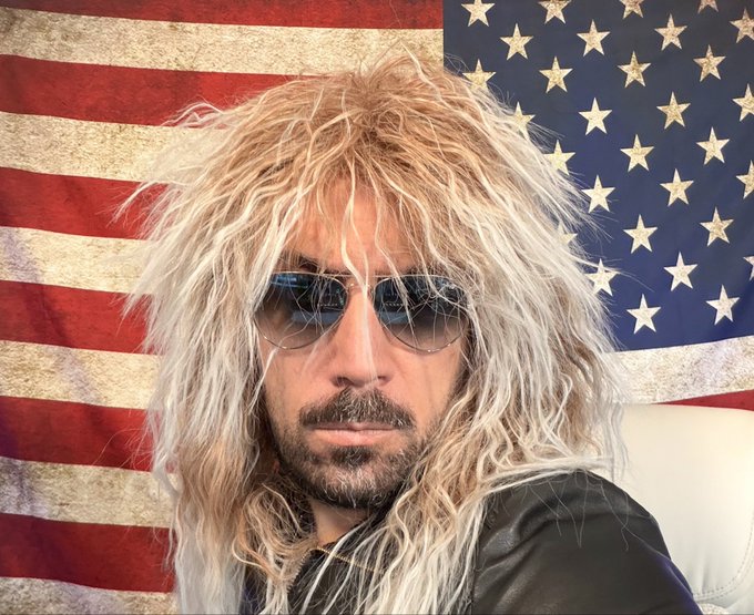 A man with long, messy blonde hair and aviator sunglasses poses in front of a distressed American flag backdrop, wearing a black shirt while seated in what appears to be a white chair. The rugged, rock-star aesthetic of the portrait complements the post's question about "Rex Liberty" potentially being the catalyst for a bull run, evoking themes of bold, market-disrupting energy.