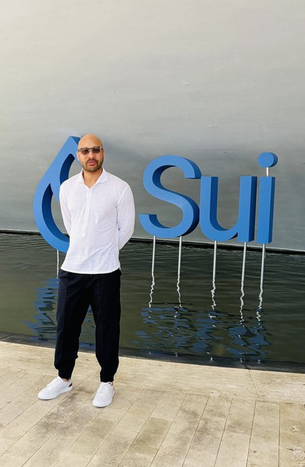 A man in a white shirt and black pants standing in front of a large blue 'Sui' logo sign by the water, wearing sunglasses.