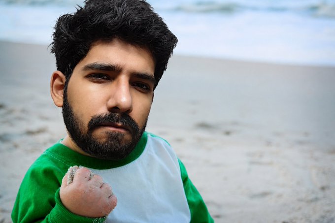 A close-up portrait of a man with dark hair and a beard, wearing a green and white shirt, holding a fist of sand on a beach.