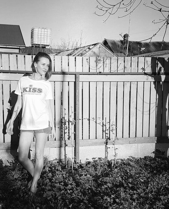 A black and white photo shows a woman wearing a KISS t-shirt and denim shorts standing barefoot in a garden next to a wooden fence.