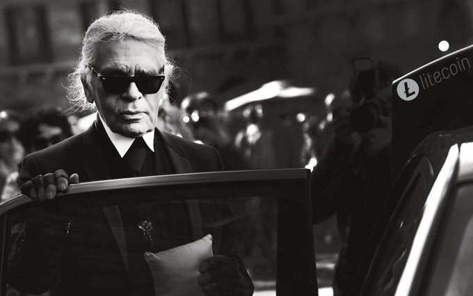 A black-and-white photo shows Karl Lagerfeld wearing his signature dark sunglasses and a black suit while exiting a car, holding papers, with a blurred crowd in the background and a "litecoin" logo visible on a vehicle to the right.