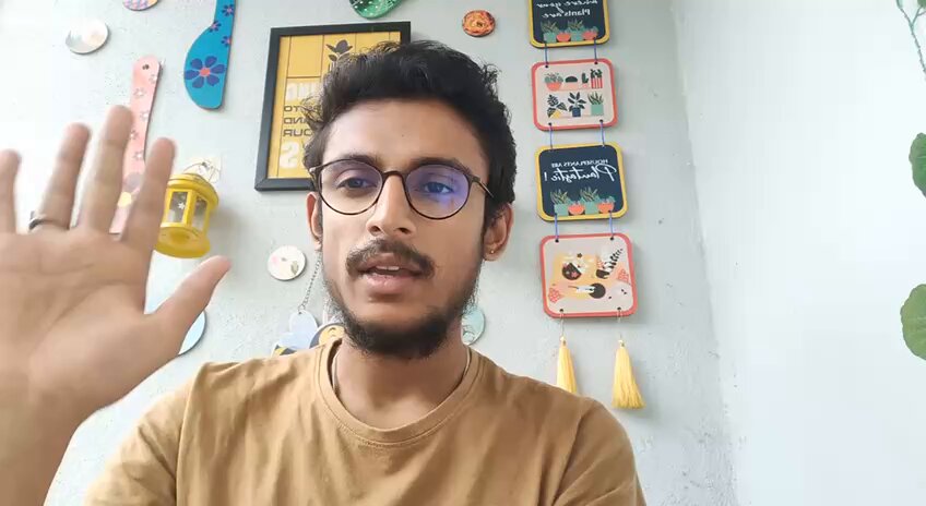 The image features a man wearing glasses and a brown shirt who appears to be making a hand gesture or waving his hand. The background of the photo includes several framed pictures, which are likely part of a wall decoration. There is also a potted plant visible in the scene.