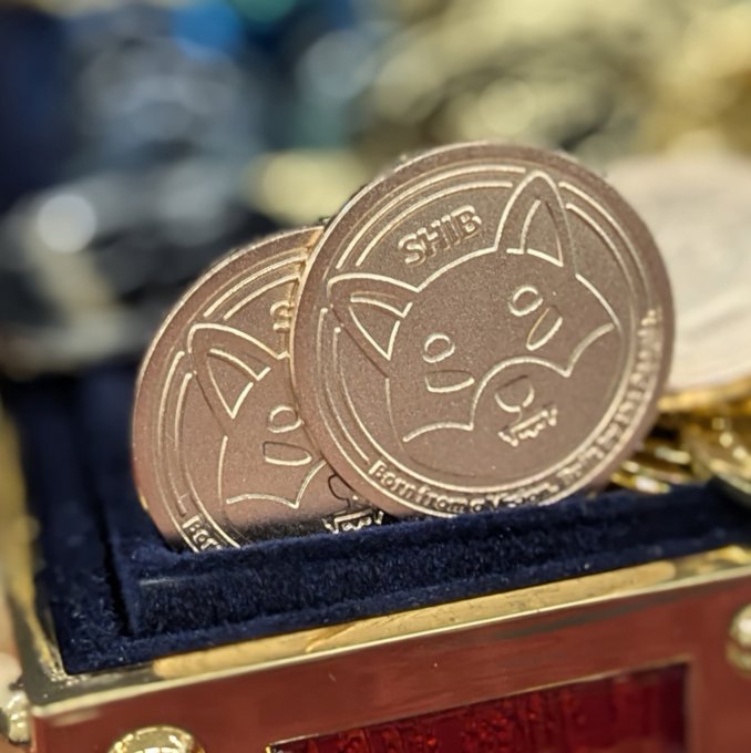 Two physical commemorative coins with a Shiba Inu design and the text 'SHIB' and 'In Crypto We Trust' rest on a blue velvet surface inside a decorative case.