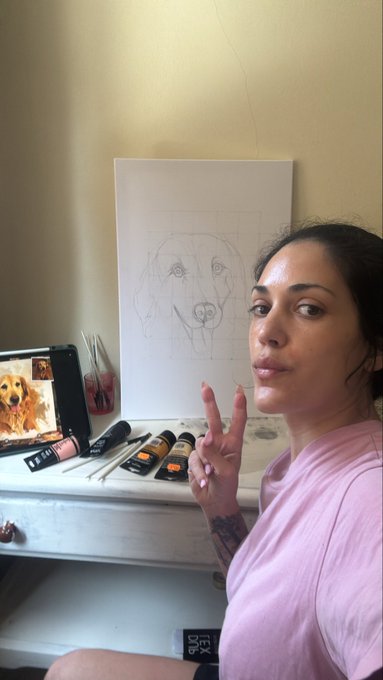 A woman in a pink shirt sits at a desk with art supplies, posing with a peace sign next to a pencil sketch of a dog on canvas.