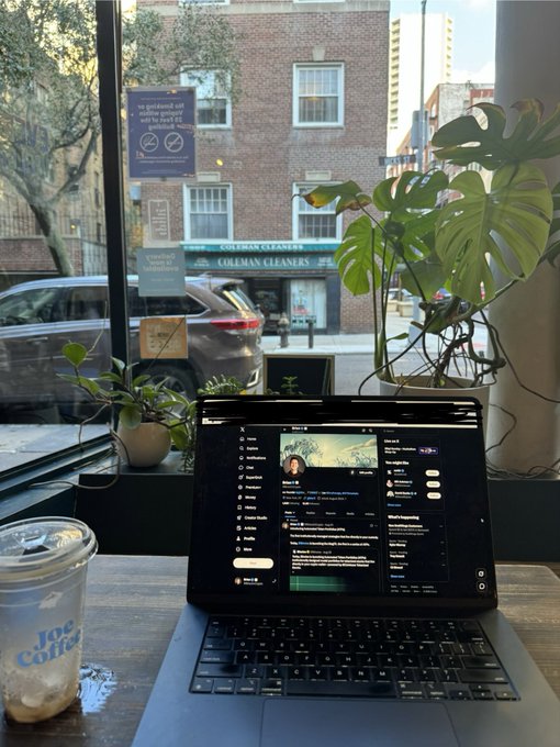 A laptop displaying a Twitter feed sits on a wooden table next to an iced coffee, with a window view showing "Coleman Cleaners" and a no-smoking sign. The image captures the quintessential work-from-cafe scene, illustrating the post's sentiment that even Sunday feels like the start of the work week.