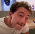A close-up image of a man with curly hair and a mustache wearing a white t-shirt, appearing to be in an indoor setting.
