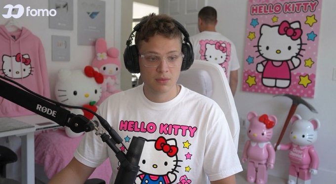 The image shows a man wearing headphones and glasses in a Hello Kitty-themed room, with a "HELLO KITTY" t-shirt, matching plush toys, and a decorative poster visible around him, while the "fomo" logo appears in the corner. The setting aligns with the post celebrating "$kitty" reaching 1,000 holders on the Fomo platform, reflecting the meme token's playful branding and community celebration.