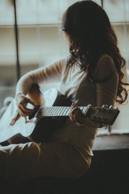 A moody, low-light photograph of a woman with long dark hair playing an acoustic guitar, her face partially obscured in shadow.