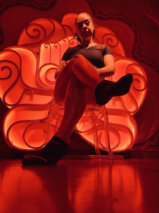 A low-angle photograph shows a person sitting on a clear plastic chair, wearing a black top, red tights, and black boots, posing against a backdrop of bright, swirling red neon lights.