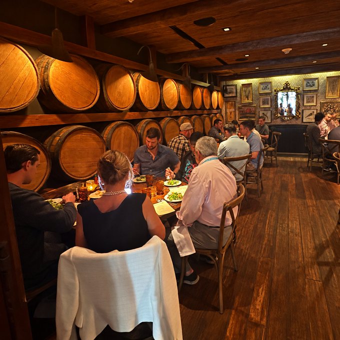 A dimly lit restaurant interior with wooden walls lined with large wine or whiskey barrels, where groups of people are seated at tables enjoying a meal.