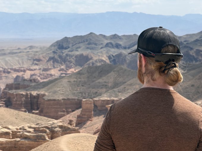 A man with a blonde bun, beard, and black cap stands overlooking a vast canyon landscape with rocky cliffs and distant mountains under a hazy sky.