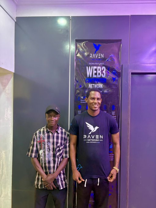 Two men standing in front of a 'RAVEN NETWORK - WEB3 POWERHOUSE' promotional banner at a building entrance.