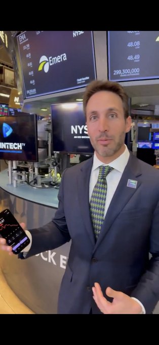 The image features a man dressed in a suit standing in front of a desk with multiple monitors, holding up a cell phone. The background includes several screens displaying stock market information. The caption reads "This is the face of the saltiest mfer on this app. Also: - President of the most negative pnl DAT - Bullposted FTX, Serum, Mango - Got ousted from his VC firm for going long Drift instead of Hyperliquid".