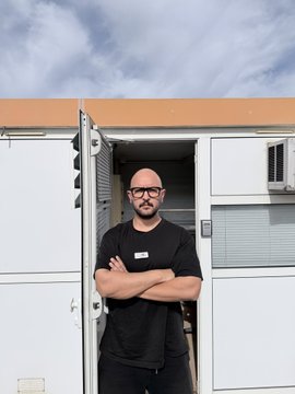 A bald man with glasses and a black t-shirt stands with crossed arms in front of an open storage unit or trailer.