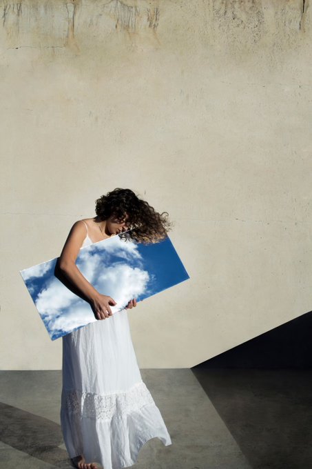 A surreal photograph of a woman in a white dress holding a rectangular board printed with a blue sky and clouds, standing against a textured beige wall.