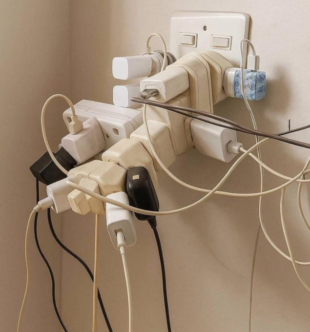 A close-up photo of a wall outlet overloaded with multiple white power adapters and tangled charging cables plugged in chaotically.