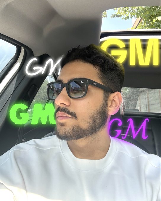 A young man with a beard and black sunglasses sits in a car, surrounded by the text "GM" displayed in four different neon styles (white, yellow, green, and purple), echoing the post's casual "GM homies" greeting.