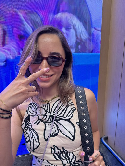 Photo of a smiling young woman wearing sunglasses and a floral top, making a peace sign with her hand in front of a large blue digital display screen.