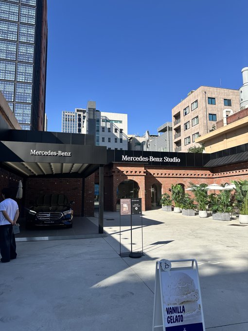 The exterior of a Mercedes-Benz Studio is shown, featuring a brick building, a black Mercedes SUV on display, and a 'Vanilla Gelato' sign in the foreground.