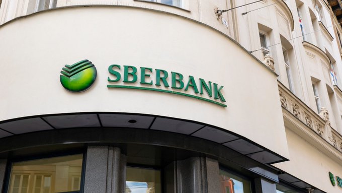 The image shows the exterior of a Sberbank branch with its distinctive green logo and name prominently displayed on the building's facade. The image visually anchors the post's news that Sberbank, Russia's largest bank, is preparing to accept cryptocurrencies like Bitcoin, Ethereum, and Tether as loan collateral under upcoming digital asset regulations.