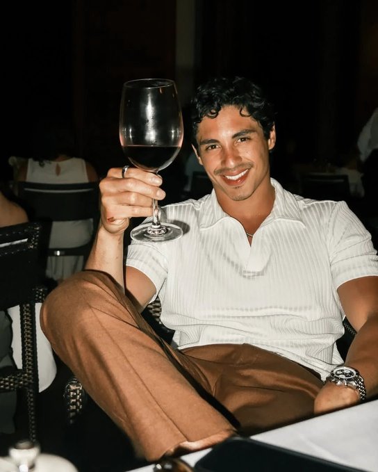A photograph of a smiling young man in a white short-sleeved knit shirt and brown pants, holding up a glass of red wine while seated at a table in a dimly lit setting.
