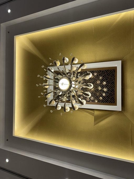 An ornate crystal chandelier illuminated with warm yellow light hangs from a recessed ceiling above a decorative panel with a dark geometric pattern.